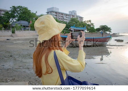 Woman standing behind and using a handheld camera to capture the sunset on the beach at sea The sea along the coast where fishing boats survive on outdoor holidays.