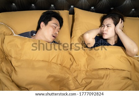 Asian man snoring with open mouth,wife is closing ears with hands,stressed by loud noise,annoying snoring of husband,risk of sleep apnea while sleeping,unhappy woman wakes up while lying in the bed Royalty-Free Stock Photo #1777092824