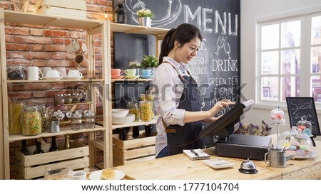 smiling coffee shop assistant using pos point of sale terminal to put in order from note paper at restaurant register. waitress lady working in counter in cafe store. young girl staff using cashbox. Royalty-Free Stock Photo #1777104704