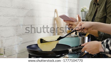 closeup two asian female cooking together in kitchen. woman hands with spatula flipping pancake in hot frying pan on top of gas stove range. friend holding mobile phone taking photo of making process
