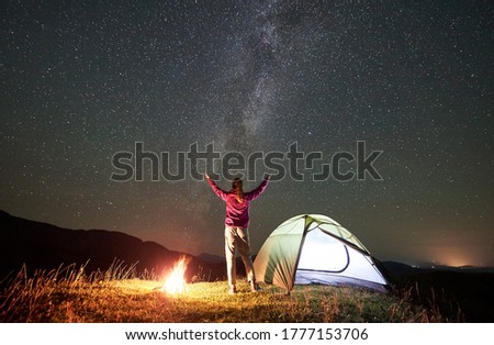 Female hiker having a rest at summer night camping beside campfire and glowing tourist tent. Back view of young woman holding hands lifting up, enjoying view of night sky full of stars and Milky way.