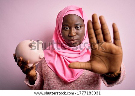 Young african american plus size woman wearing muslim hijab holding pink piggy bank with open hand doing stop sign with serious and confident expression, defense gesture