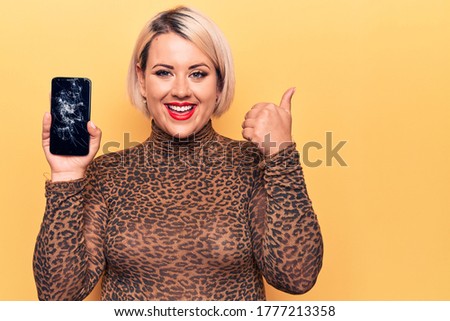 Young beautiful blonde plus size woman holding broken smartphone showing cracked screen smiling happy and positive, thumb up doing excellent and approval sign