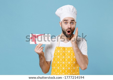 Shocked young bearded male chef or cook baker man in apron white t-shirt toque chefs hat isolated on blue background. Cooking food concept. Mock up copy space. Hold gift certificate put hand on cheek