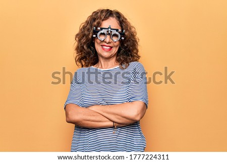 Middle age beautiful woman controlling vision using optometry glasses over yellow background happy face smiling with crossed arms looking at the camera. Positive person.