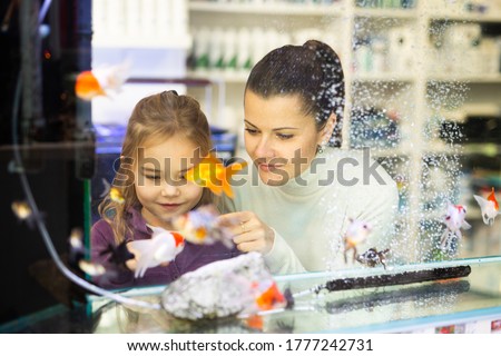 Interested little girl with smiling young mother standing behind glass aquarium in pet store choosing exotic fish for home fish tank

