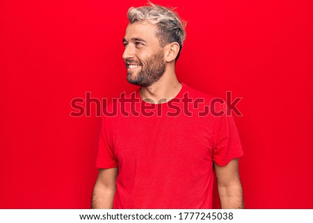 Young handsome blond man wearing casual red t-shirt standing over isolated red background looking to side, relax profile pose with natural face and confident smile.