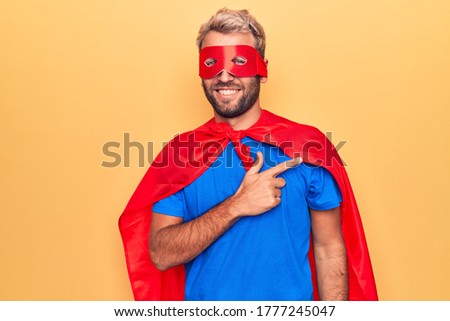 Handsome blond man wearing super hero costume with mask and cape over yellow background smiling cheerful pointing with hand and finger up to the side