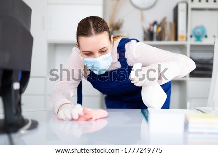 Woman professional cleaner in protective medical mask cleaning desk with detergents in office Royalty-Free Stock Photo #1777249775