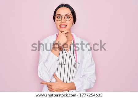 Beautiful doctor woman with blue eyes wearing coat and stethoscope over pink background looking confident at the camera smiling with crossed arms and hand raised on chin. Thinking positive.