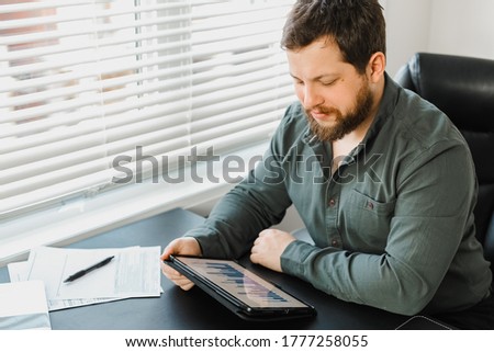 Serious male economist using digital tablet and working with documents at office. Concept of deadline, modern technology and business. Adult bearded man browsing by gadget.
