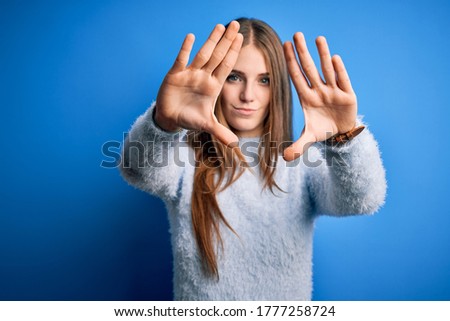 Young beautiful redhead woman wearing casual sweater over isolated blue background doing frame using hands palms and fingers, camera perspective