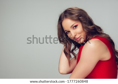 Beautiful young woman with long healthy curly hair and bright make up wearing red dress isolated on grey studio backgroud. Beauty and fashion, hair and skin care, treatment concept. Well-kept girl.