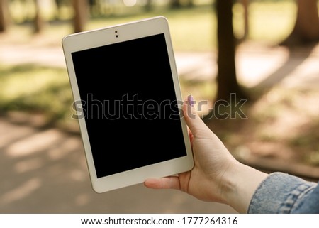 Hand woman holding white tablet pc in the city park in sunny weather, copy space and mock up. Cropped shot of female showing picture in the internet. 5G network using concept.