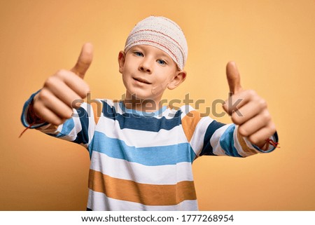 Young little caucasian kid injured wearing medical bandage on head over yellow background approving doing positive gesture with hand, thumbs up smiling and happy for success. Winner gesture.