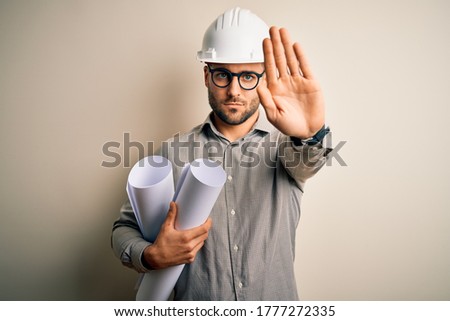 Young architect man wearing contractor helmet holding project paper plan over isolated background with open hand doing stop sign with serious and confident expression, defense gesture