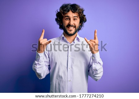 Young handsome business man with beard wearing shirt standing over purple background shouting with crazy expression doing rock symbol with hands up. Music star. Heavy music concept.
