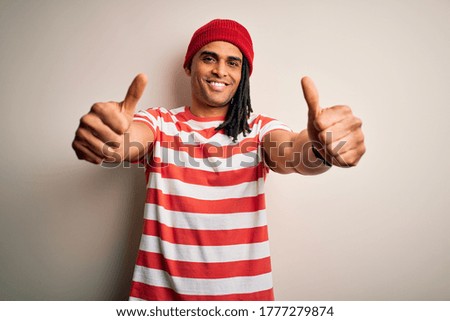 Young handsome african american man with dreadlocks wearing striped t-shirt and wool hat approving doing positive gesture with hand, thumbs up smiling and happy for success. Winner gesture.