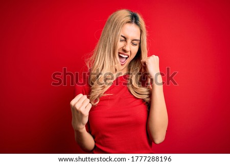 Young beautiful blonde woman wearing casual t-shirt standing over isolated red background celebrating surprised and amazed for success with arms raised and eyes closed. Winner concept.