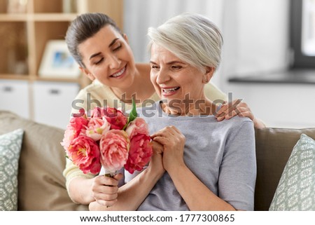 family, mother's day and birthday concept - happy smiling adult daughter giving flowers to her senior mother at home