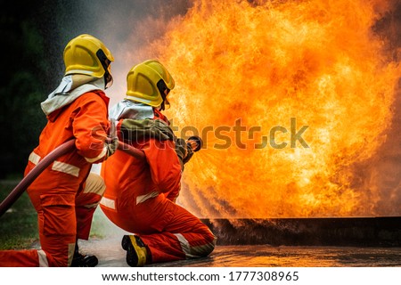 Two brave firefighter in fire suit on rescue duty using water from hose extinguishing fighting with big crackle fire flames inside burning premises. Fireman spraying high pressure water fight a fire.