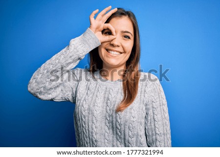 Beautiful young woman wearing casual wool sweater standing over blue isolated background doing ok gesture with hand smiling, eye looking through fingers with happy face.