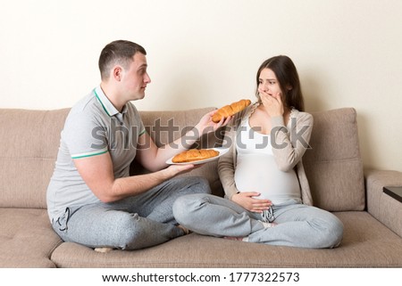 Husband offers croissants to his pregnant wife but she refuses and makes stop gesture because she feels sick. Feeling bad during pregnancy concept.