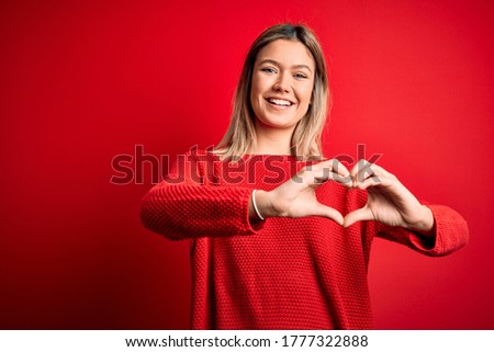 Young beautiful blonde  wearing casual sweater over red isolated background smiling in love doing heart symbol shape with hands. Romantic concept.