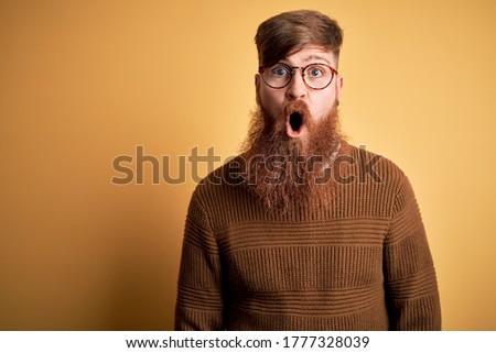 Handsome Irish redhead man with beard wearing glasses and winter sweater over yellow background afraid and shocked with surprise expression, fear and excited face.