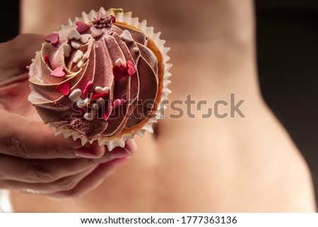 Female hands hold a sweet, appetizing cupcake against the background of a flat, toned female belly, and a thin waist. Unhealthy small amounts of sweet foods can be healthy, not harmful.