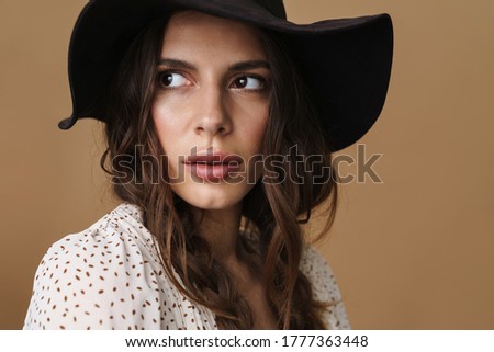Photo of brunette young woman in hat posing and looking aside isolated over beige background