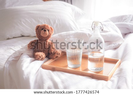 A teddy bear on the bed with a bottle and a mug in a wooden tray