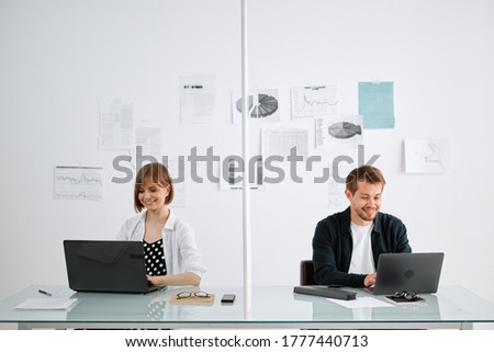 Pretty smiling girl and guy work at computers in office at a safe distance from each other