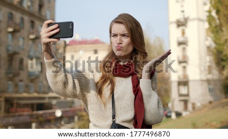Portrait of flirty girl taking selfie photo on mobile phone outdoors. Closeup hipster woman making faces on smartphone camera outside. Cute female person grimacing for cellphone camera on street