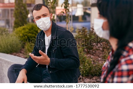 Young guy in medical mask gesticulating and talking with woman while sitting on border and keeping social distance during date on city street Royalty-Free Stock Photo #1777530092