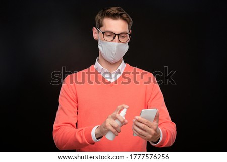 Young man in mask and eyeglasses disinfecting smartphone with sanitizer against black background, coronavirus concept