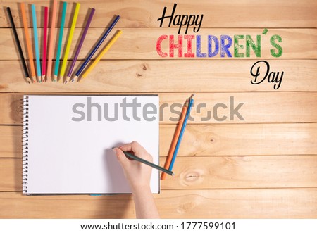 Child holding a pencil on top of a blank sheet surrounded by colored pencils. Text: Happy Childrens Day