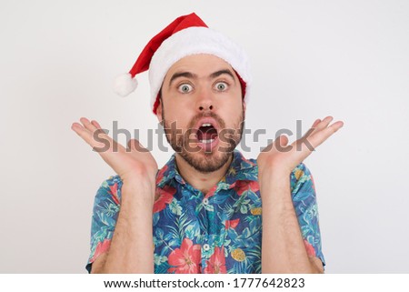Surprised terrified  Young caucasian man wearing hawaiian shirt and Christmas hat over isolated white background Gestures with uncertainty, stares at camera, puzzled as doesn't know answer 