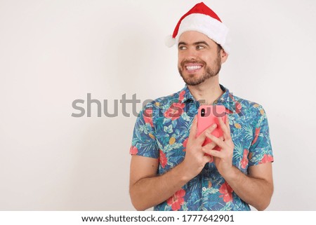 Young caucasian man wearing hawaiian shirt and Christmas hat over isolated white background hold telephone hands read good youth news look empty space advert 