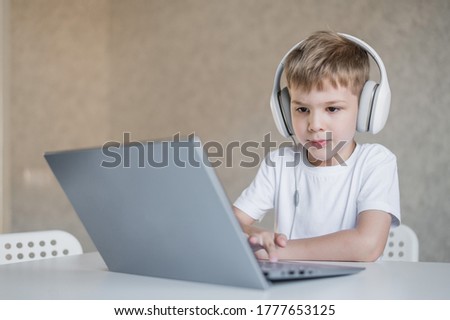 Cute little boy is watching cartoons or a movie on a laptop. Caucasian child sits at a table at home and listens to music on headphones.