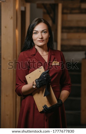 A female restaurant manager in black disposable medical gloves is posing holding a menu in a restaurant. A kind cafe owner in a ruby dress.