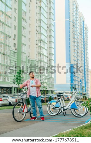 Adult man stands in the yard against the backdrop of a high-rise building with a rented city bike and smiling looking to the side. Eco transport for a walk around the town.