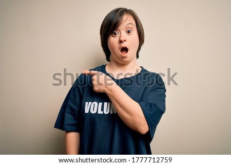 Young down syndrome volunteer woman wearing social care charity t-shirt Surprised pointing with finger to the side, open mouth amazed expression.