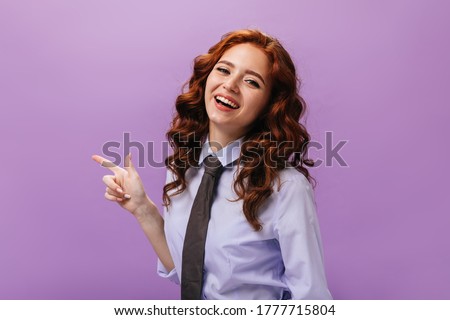 Lady in blue shirt pointing to place for text on purple background. Curly beautiful woman in tie and lilac shirt smiling