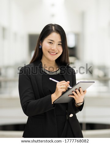 Portrait of a young cheerful businesswoman surfing social network on digital tablet in front of office during break. Asian business woman standing in office building. ฺBusiness stock photo.
