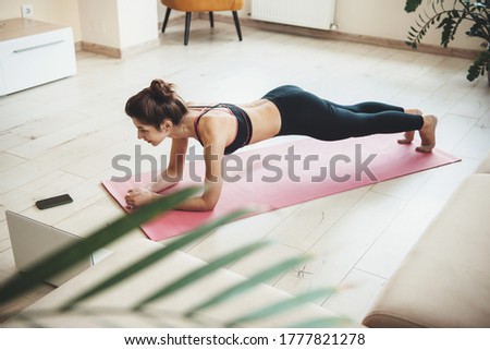 Caucasian brunette woman planking at home during the lockdown wearing sportswear and using a laptop with phone