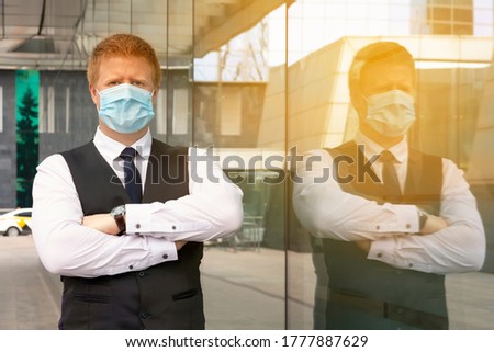Adult caucasian man wearing protective mask stands with crossed arms near the window of office building. The man is wearing a business uniform. Theme of work during the coronavirus pandemic.
