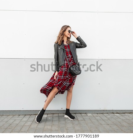 Joyful hipster woman in fashion sunglasses in fashionable casual beautiful red-black clothes in stylish sneakers with leather bag posing near wall in city. Smiling woman model on street.Trendy look.