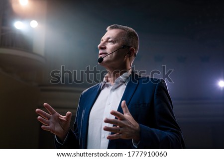Portrait of men speaking through a microphone in dark conference hall. Person says about marketing and management for successful sales to college students indoors closeup. Professional ideas, politics Royalty-Free Stock Photo #1777910060