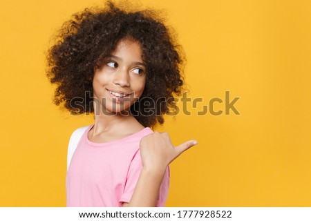 Side view of smiling little african american kid girl 12-13 years old in pink t-shirt isolated on yellow wall background studio. Childhood lifestyleconcept. Mock up copy space. Pointing thumb aside Royalty-Free Stock Photo #1777928522